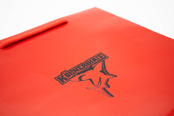 Sac en papier rouge avec logo noir des Kölner Haie et poignée en cordelette